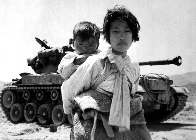 With her brother on her back a war weary Korean girl tiredly trudges by a stalled M-26 tank, at Haengju, Korea. June 9, 1951.