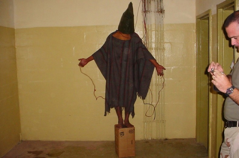 An unidentified detainee standing on a box with a bag on his head and wires attached.