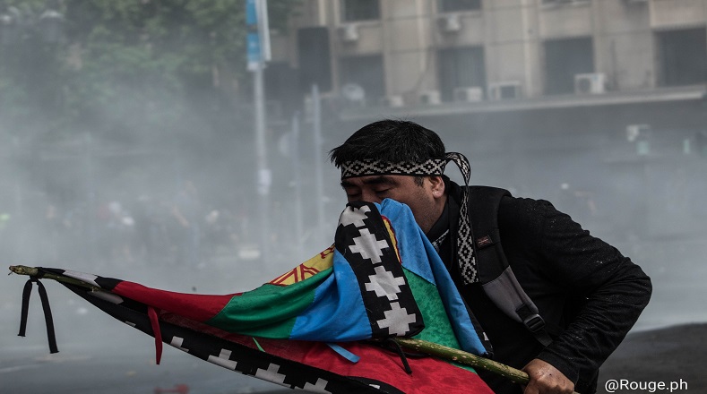 Muestra a un miembro de la comunidad mapuche (pueblo originario de la Araucanía) siendo afectado por los gases químicos disparados por fuerzas especiales en una manifestación pacífica, este usa la bandera de su pueblo para cubrirse el rostro y así intentar que el efecto no sea tan dañino.